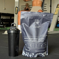 Coconut flavoured Plant protein powder, shown in a gym whilst a woman is using the chest press machine.